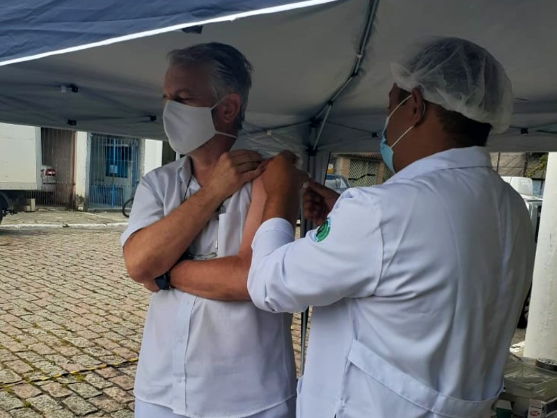 Profissional da saúde recebe a vacina no braço embaixo de uma tenda. Quem aplica é outro profissional da saúde. Os dois estão de roupas brancas e máscara